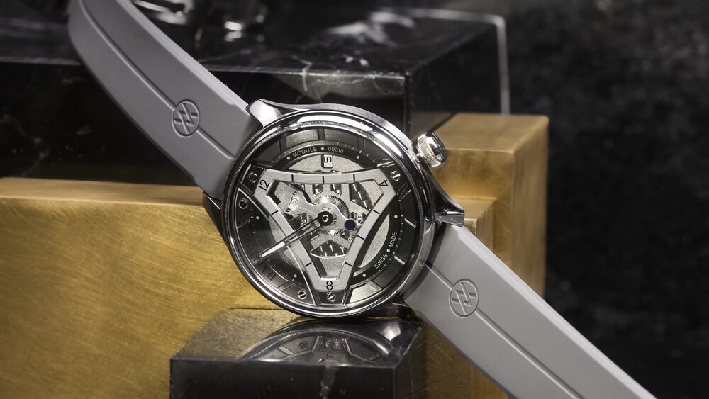 A silver watch with a geometric dial and grey strap, placed on a reflective surface.