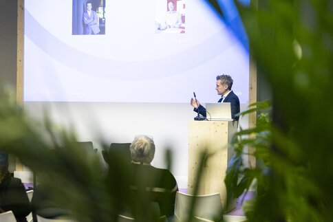 A man is standing at a lectern, with a large screen behind him. He is giving a talk to a small audience. The image is framed by green plants.