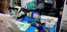 Woman in a lab coat sits at a table with a microscope, monitors and soldering irons during productronica. Other people are standing in the background.