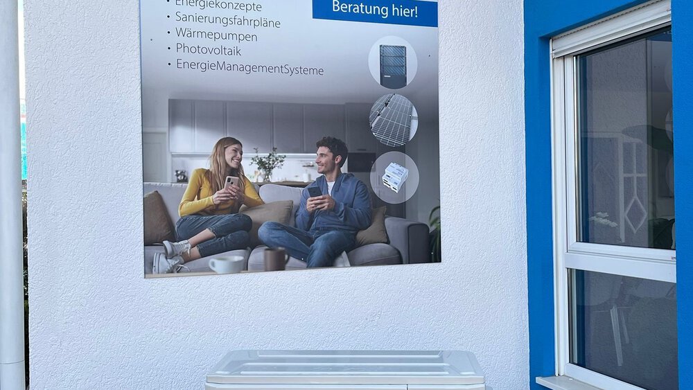 Ein Wandschild wirbt für Zewotherm, während ein Paar auf einer Couch sitzt; darunter befindet sich ein Klimagerät für den Außenbereich.