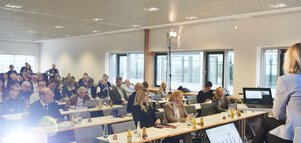 Menschen sitzen in mehreren Reihen an Tischen in einem Konferenzraum und hören sich eine Präsentation an.