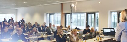 Menschen sitzen in mehreren Reihen an Tischen in einem Konferenzraum und hören sich eine Präsentation an.