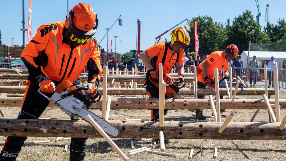 Drei Männer in orangefarbener Ausrüstung schneiden bei der INTERFORST im Freien mit Kettensägen Holzstücke ab, die in liegende Baumstämme gesteckt wurden.