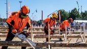 Drei Männer in orangefarbener Ausrüstung schneiden bei der INTERFORST im Freien mit Kettensägen Holzstücke ab, die in liegende Baumstämme gesteckt wurden.