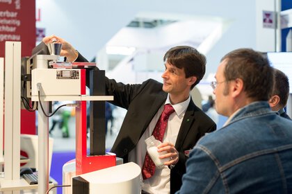 Auf der Messe analytica gibt ein Mann im Anzug ein weißes Pulver in ein Analysegerät und demonstriert die Funktion des Geräts zwei Besuchern.