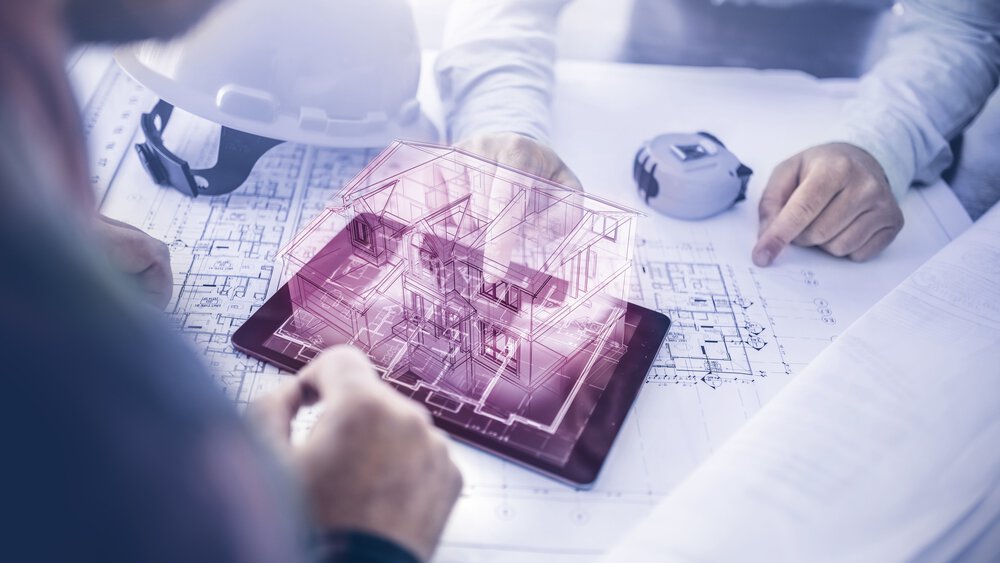 Two people are sitting at a table with construction plans. Between them is a tablet with the purple outline of a 3D house protruding from it.
