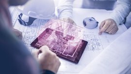 Two people are sitting at a table with construction plans. Between them is a tablet with the purple outline of a 3D house protruding from it.