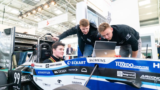 Ein Mann sitzt in einem Rennwagen der auf einem Messestand ausgestellt ist. Zwei weitere Männer beugen sich über das Fahrzeug und inspizieren etwas auf einem Tablet.