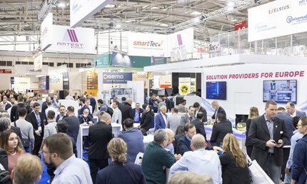 Ein sehr gut besuchter Bereich der productronica, in dem sich Besucher in Gruppen austauschen, während über ihren Köpfen Banner mit Logos der Aussteller prangen.