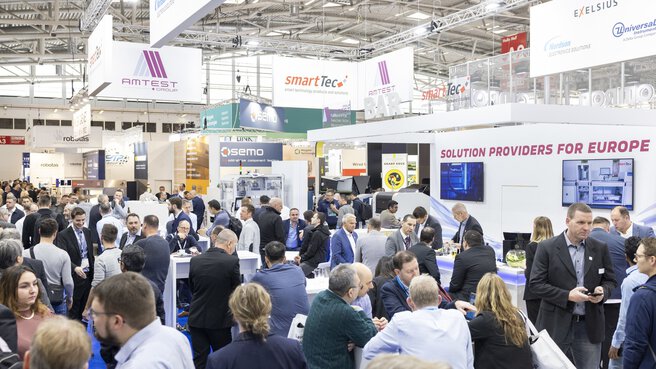 Ein gut besuchter Bereich der productronica, in dem sich Besucher in Gruppen austauschen, während über ihren Köpfen Banner mit Logos der Aussteller prangen.