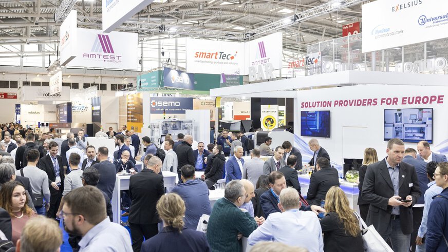 Ein sehr gut besuchter Bereich der productronica, in dem sich Besucher in Gruppen austauschen, während über ihren Köpfen Banner mit Logos der Aussteller prangen.