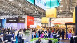 Exhibition stands of major companies and numerous visitors engaged in lively conversations at EXPO REAL 2026 in Munich, creating a vibrant atmosphere.