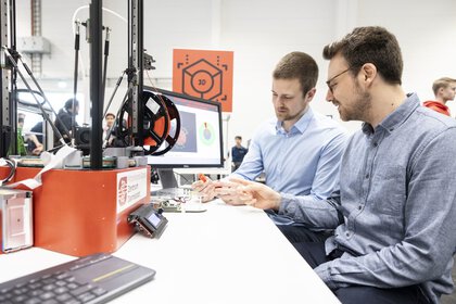 Zwei Männer sitzen an einem Tisch, auf dem ein 3D-Drucker und ein eingeschalteter Monitor stehen. Sie betrachten ein kleines fertiges Werkstück des 3D-Druckers.