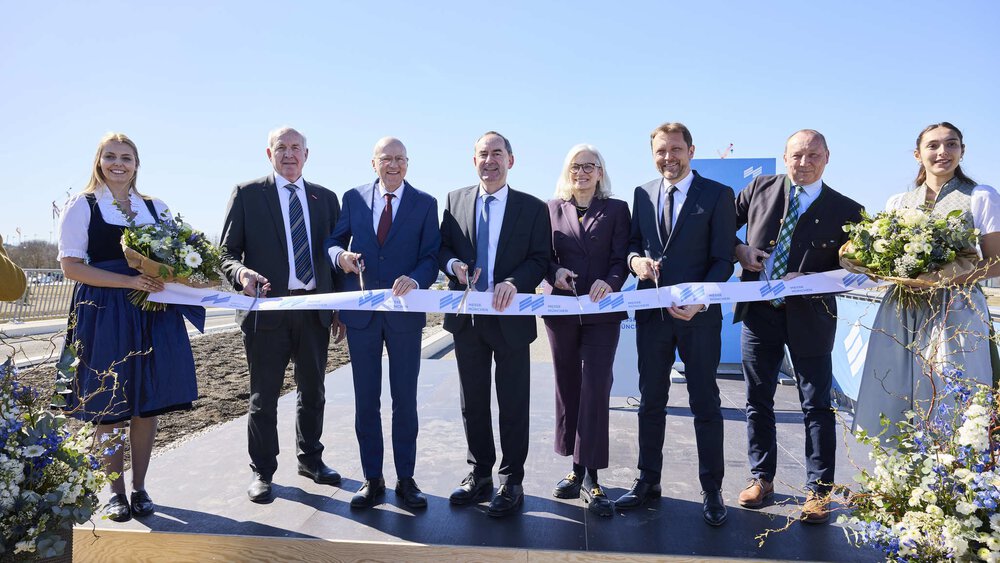 Acht Personen bei einer feierlichen Eröffnungszeremonie mit Blumen und blauem Himmel im Hintergrund.