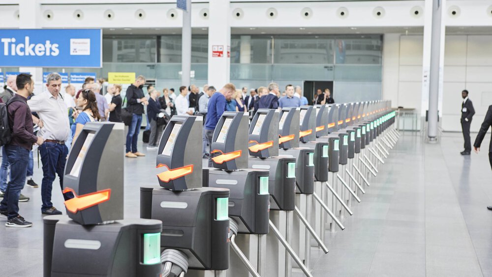 A row of electronic turnstiles at an airport or train station, with people waiting in the background.