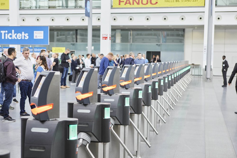 A row of electronic turnstiles at an airport or train station, with people waiting in the background.
