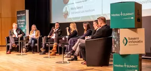 Seven people sit on a stage and discuss at the MedtecSUMMIT event; a screen and banners can be seen in the background.