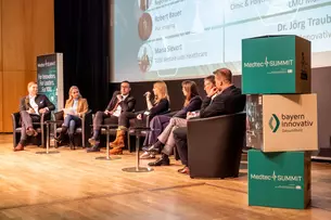 Seven people sit on a stage and discuss at the MedtecSUMMIT event; a screen and banners can be seen in the background.