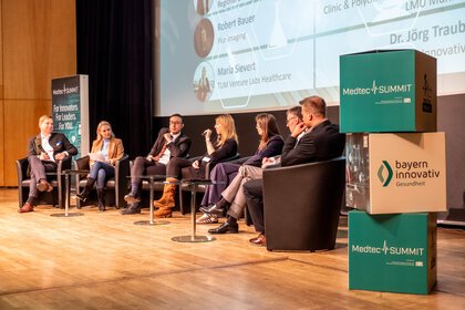 Sieben Personen sitzen auf einer Bühne und diskutieren bei der Veranstaltung MedtecSUMMIT, im Hintergrund sind ein Bildschirm und Banner zu sehen.