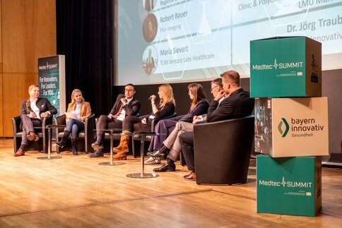 Seven people sit on a stage and discuss at the MedtecSUMMIT event; a screen and banners can be seen in the background.