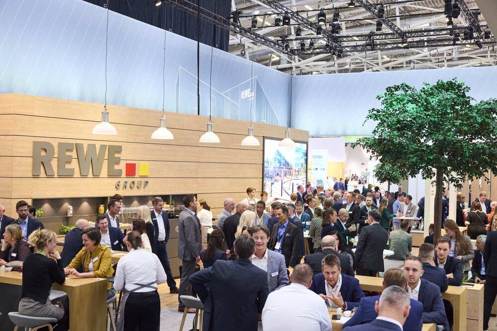 Many people make contacts at a REWE Group stand in a large, modern exhibition hall.