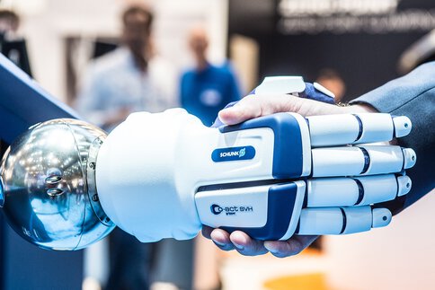 Nahaufnahme von Roboterhand und Menschenhand geben sich die Hand. Im Hintergund sieht man weitere Personen.