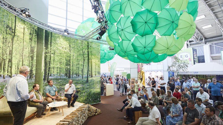A seated audience watches a panel discussion on a low stage with four people. Green umbrellas have been hung from the ceiling to form large spheres.