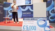 Person speaking on stage at an investor summit event with a large prize check for €5,000.