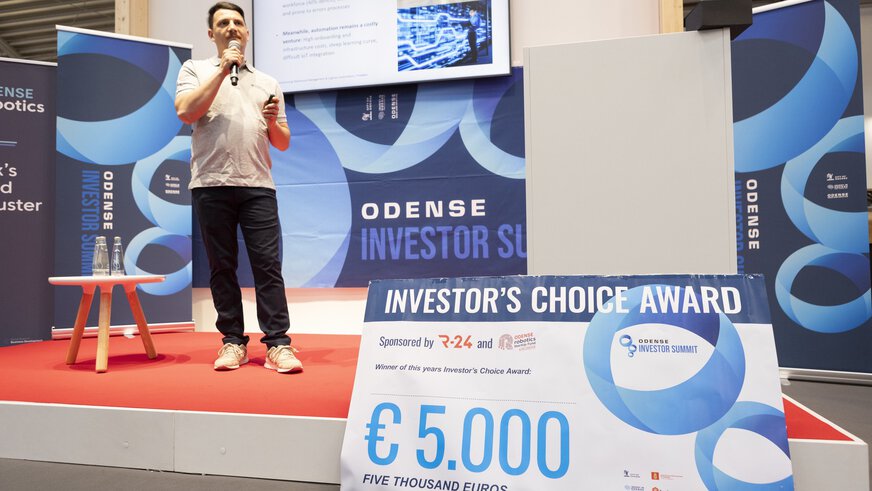 Person speaking on stage at an investor summit event with a large prize check for €5,000.