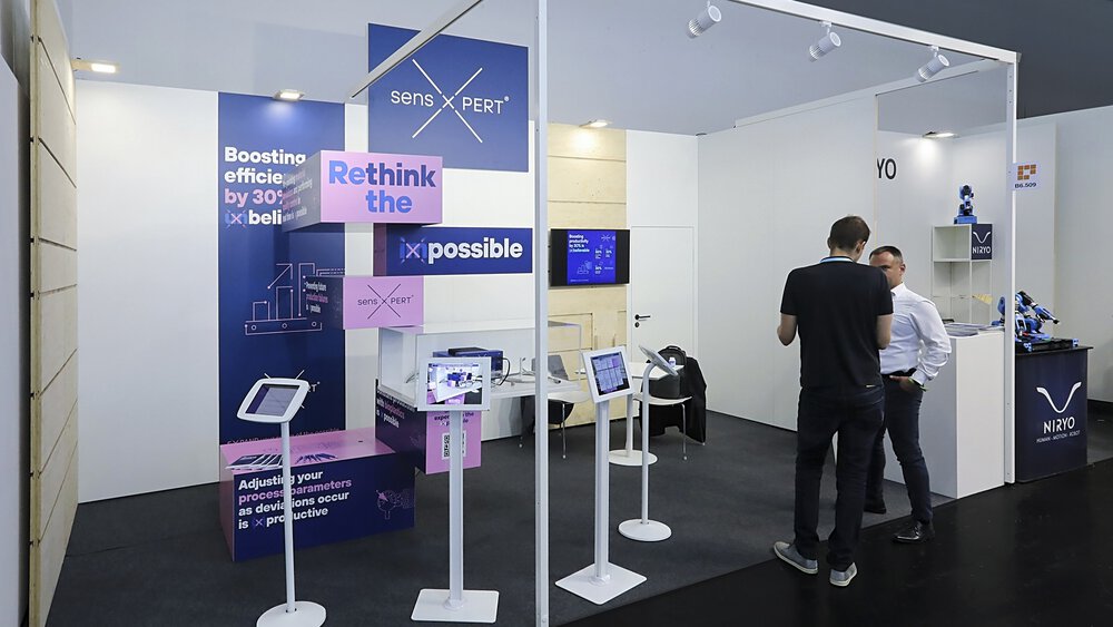 Messestand mit technischen Displays, Bildschirmen und zwei interagierenden Personen. Auf den Schildern steht 