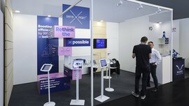 Messestand mit technischen Displays, Bildschirmen und zwei interagierenden Personen. Auf den Schildern steht 