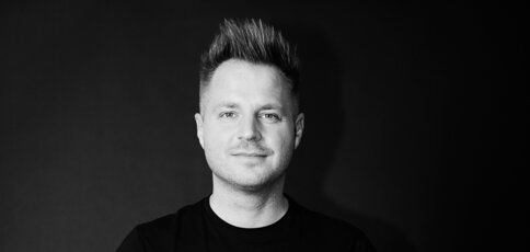 Lächelnder Mann mit kurzen hochstehenden Haaren trägt ein schwarzes Hemd, vor dunklem Hintergrund.