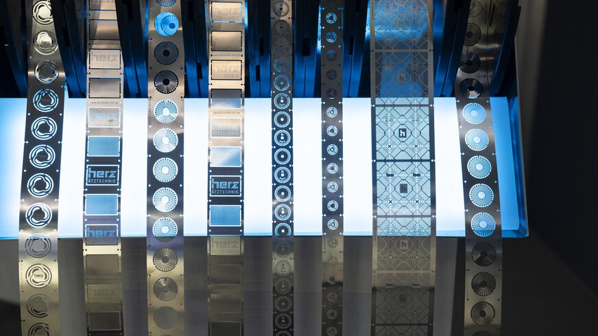 Lange Blechstreifen mit ausgestanzten Logos hängen herab, dahinter ein blau leuchtendes Display.