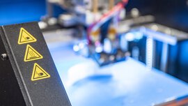 Three triangular, yellow warning stickers on the housing of a machine that is only blurred in the background.