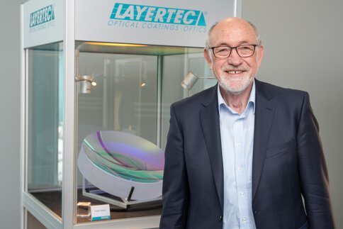 Lächelnder Mann mit weißem Bart, im Anzug neben einer Vitrine mit dem Logo 
