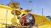 A drummer plays in a yellow excavator shovel.