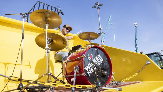 A drummer plays in a yellow excavator shovel.