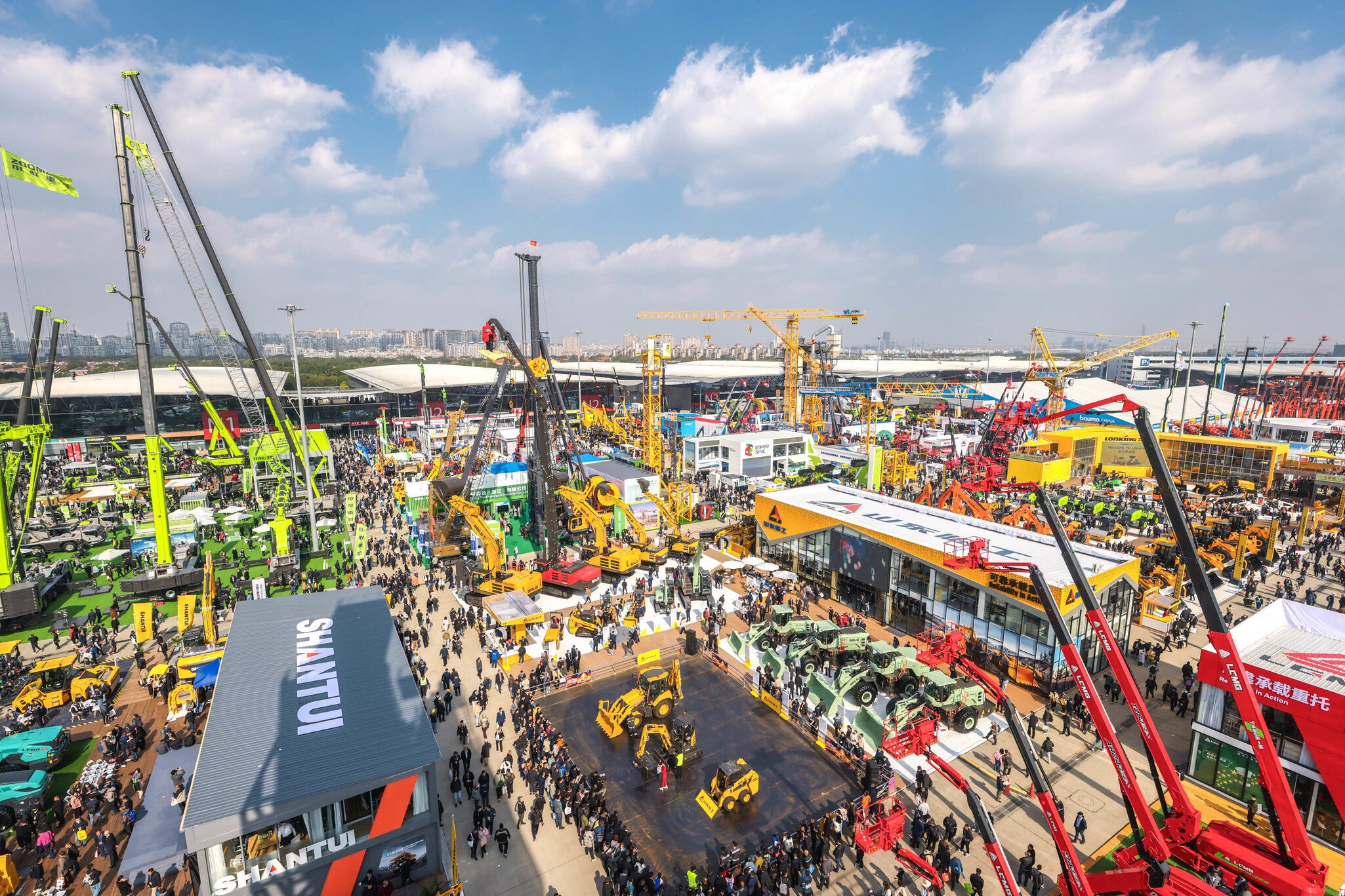 A large outdoor construction machinery exhibition with crowds of people, cranes and machinery under a partly cloudy sky.