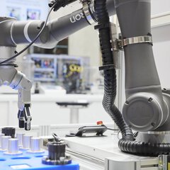 Ein grauer Roboterarm hebt mit seinen Greifern metallene Zylinderstempel aus einer blauen Form. Im Hintergrund sind weitere Maschinen und Displays zu sehen.