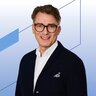 Ein lächelnder Mann mit braunen Haaren und leichtem Bart trägt eine auffällige dunkle Brille, ein weißes Hemd und ein dunklen Cardigan mit Einstecktuch.