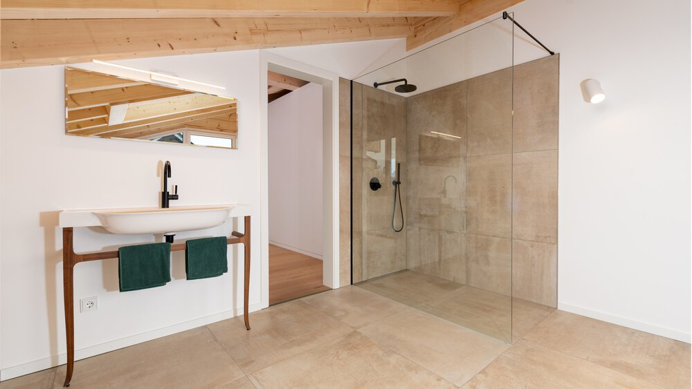 Modernes Badezimmer mit Holzdeckenbalken, durchsichtiger Glasdusche, freistehendem ovalen Waschbecken, eckigem Spiegel und beigem Fliesenboden.