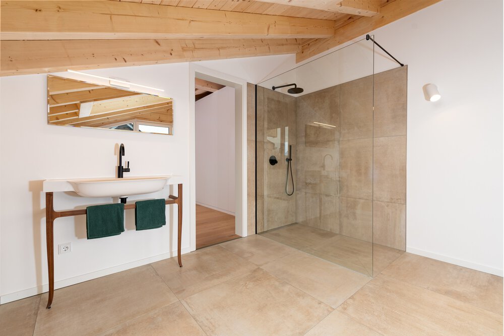Modernes Badezimmer mit Holzdeckenbalken, durchsichtiger Glasdusche, freistehendem ovalen Waschbecken, eckigem Spiegel und beigem Fliesenboden.