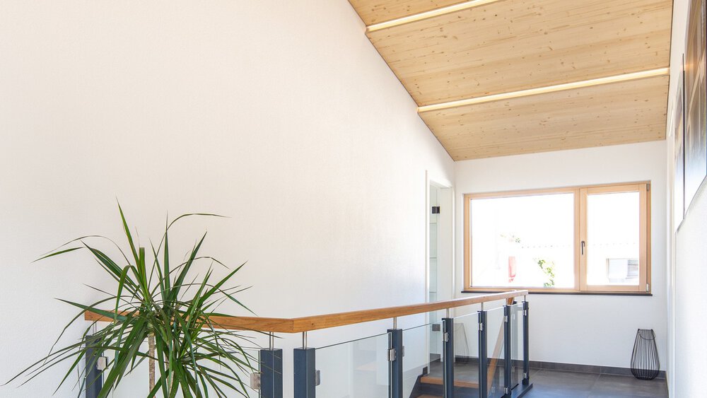 Heller Flur mit Holzdecke, großem Fenster, Pflanzen und Treppe mit schwarzem Geländer.