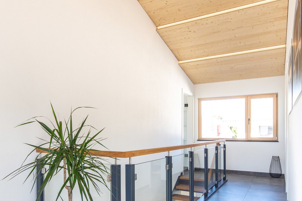 Heller Flur mit Holzdecke, großem Fenster, Pflanzen und Treppe mit schwarzem Geländer.