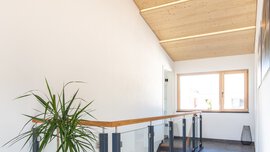 Heller Flur mit Holzdecke, großem Fenster, Pflanzen und Treppe mit schwarzem Geländer.