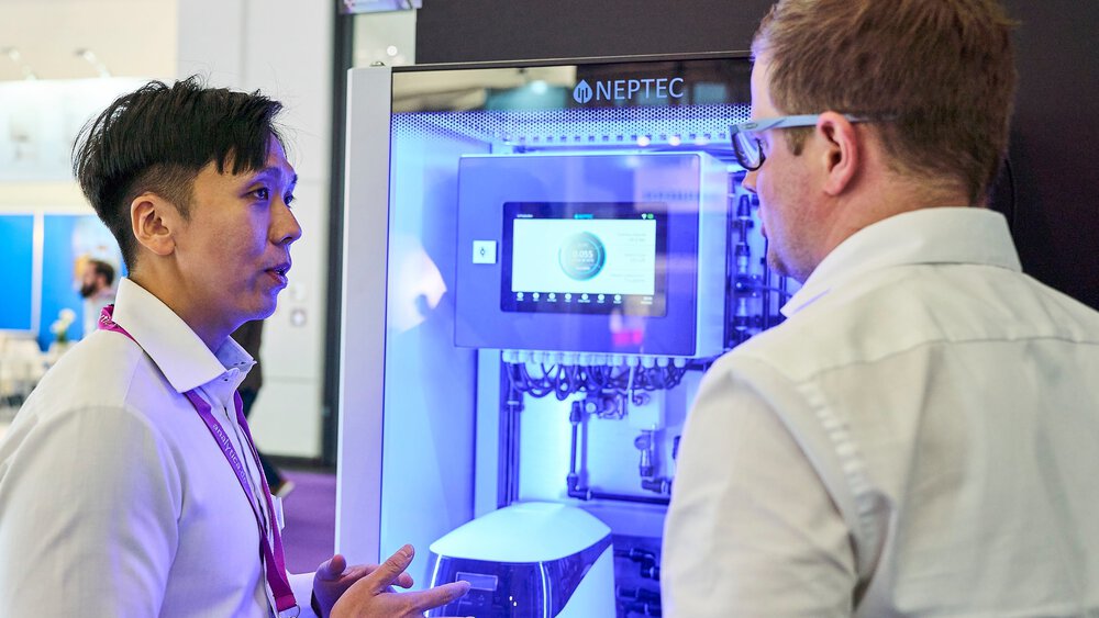 Zwei Fachbesucher sprechen vor einem beleuchteten Analysegerät im Stand.