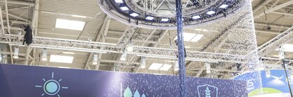 Large mast on a trade fair stand with a circular ring of LED lights in front of which a curtain of water droplets trickles down.