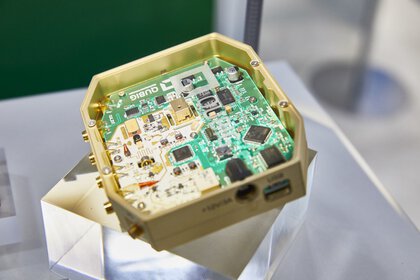 Electronic circuit board in a gold-colored housing, presented on a transparent pedestal.