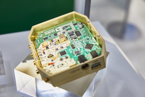 Electronic circuit board in a gold-colored housing, presented on a transparent pedestal.