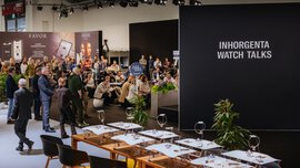 View from outside onto a stage in front of which numerous visitors are standing and sitting, listening to a lecture. Jewellery tables are set up in the foreground.
